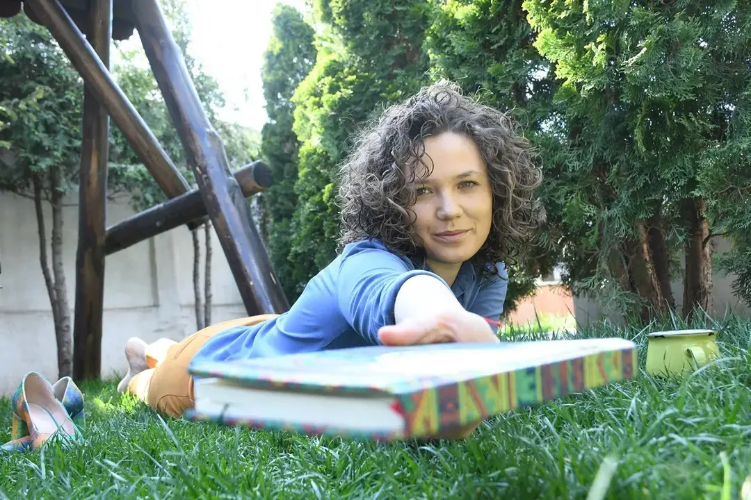 Alina Diaconu, autoare de cărți pentru copii, întinde o carte multicoloră privind în obiectiv, cu o grădină verde în fundal.